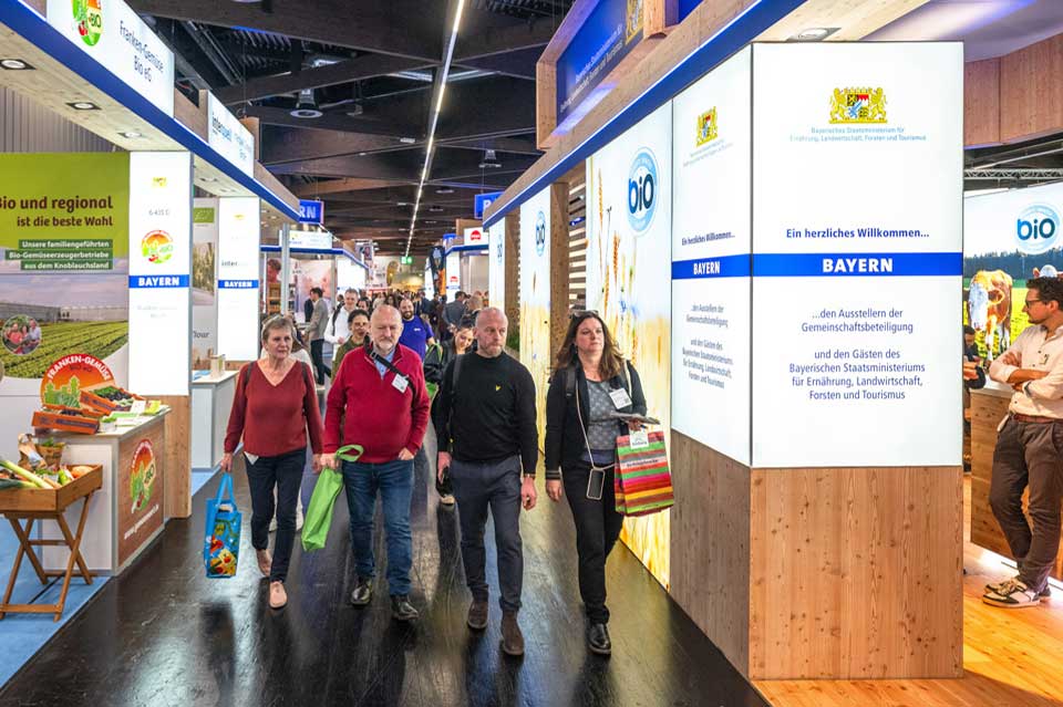 Besucher:innen gehen durch einen breiten Gang auf einer Fachmesse. Links und rechts stehen Messestände mit der Aufschrift „BAYERN“ und Informationsflächen zu Bio- und Regionalthemen. Die Stände sind aus Holz gestaltet, mit beleuchteten Tafeln und Produktpräsentationen. Mehrere Personen tragen Taschen und kommen an den Ständen vorbei ins Gespräch.