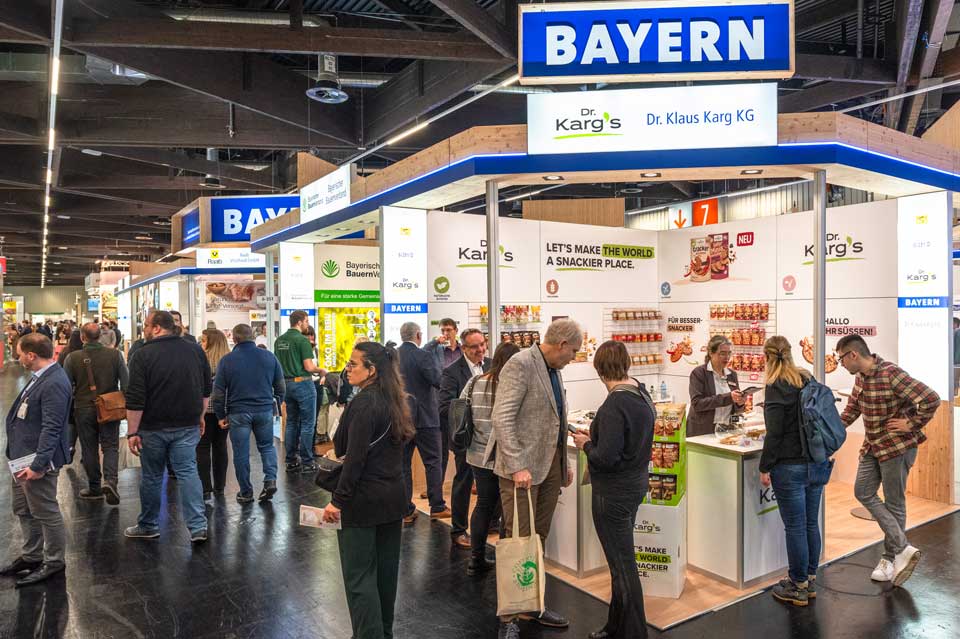 Großer Messestand mit der Aufschrift „BAYERN“ auf einer Fachmesse. Mehrere Besucher:innen stehen im Gespräch an Theken und Informationsflächen. Der Stand ist aus Holz gestaltet und präsentiert bayerische Lebensmittelhersteller, darunter Backwaren und Bio-Produkte.