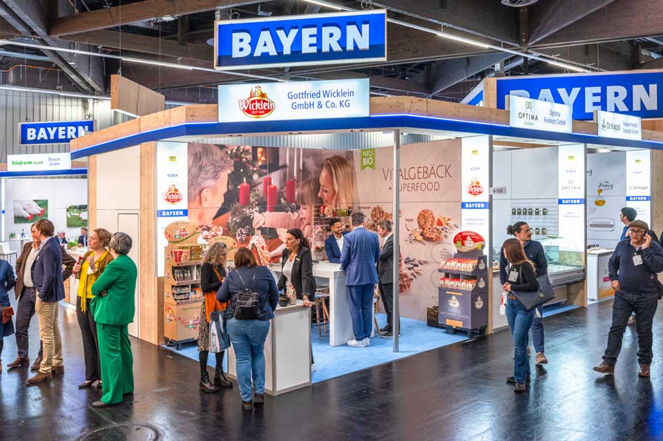 Großer Messestand mit der Aufschrift „BAYERN“ auf einer Fachmesse. Mehrere Besucher:innen stehen im Gespräch an Theken und Informationsflächen. Der Stand ist aus Holz gestaltet und präsentiert bayerische Lebensmittelhersteller, darunter Backwaren und Bio-Produkte. Im Hintergrund sind Produktregale, Kühltheken und großformatige Bildmotive zu sehen.